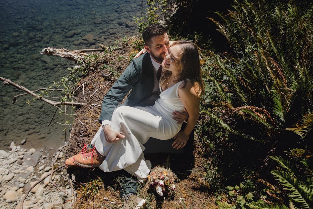 A couple in formal attire embraces while standing in a lush forest setting near a rocky stream.