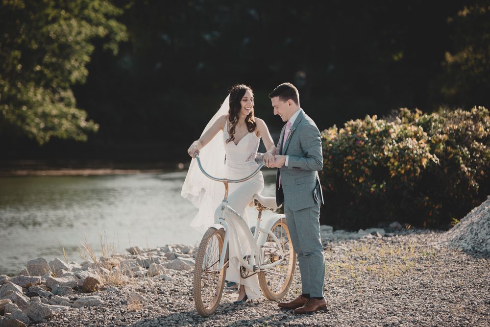 A couple in wedding attire share a romantic moment with a vintage bicycle by a lakeside during golden hour.