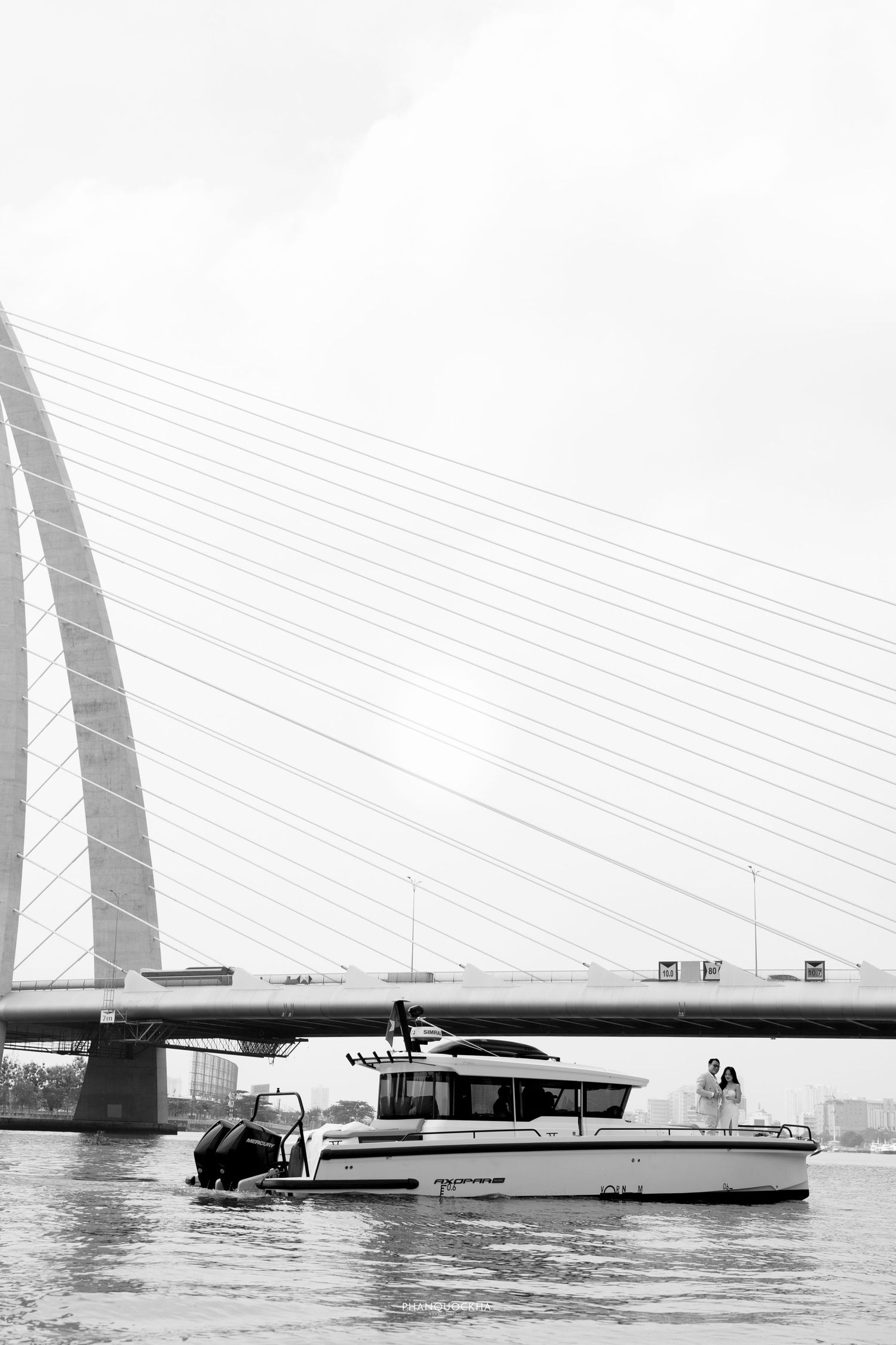 Black and white wedding photo of couple standing on a yacht beneath a modern cable-stayed bridge.