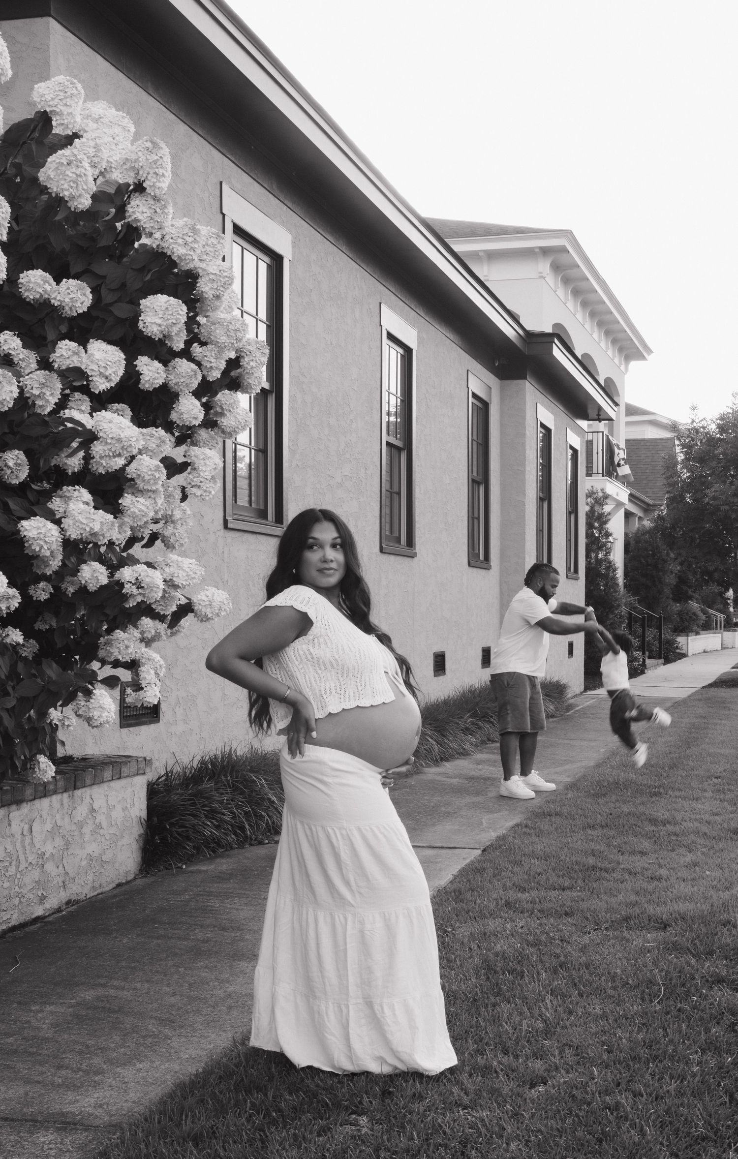 An expectant mother in a white dress poses in front of a house with blooming flowers.