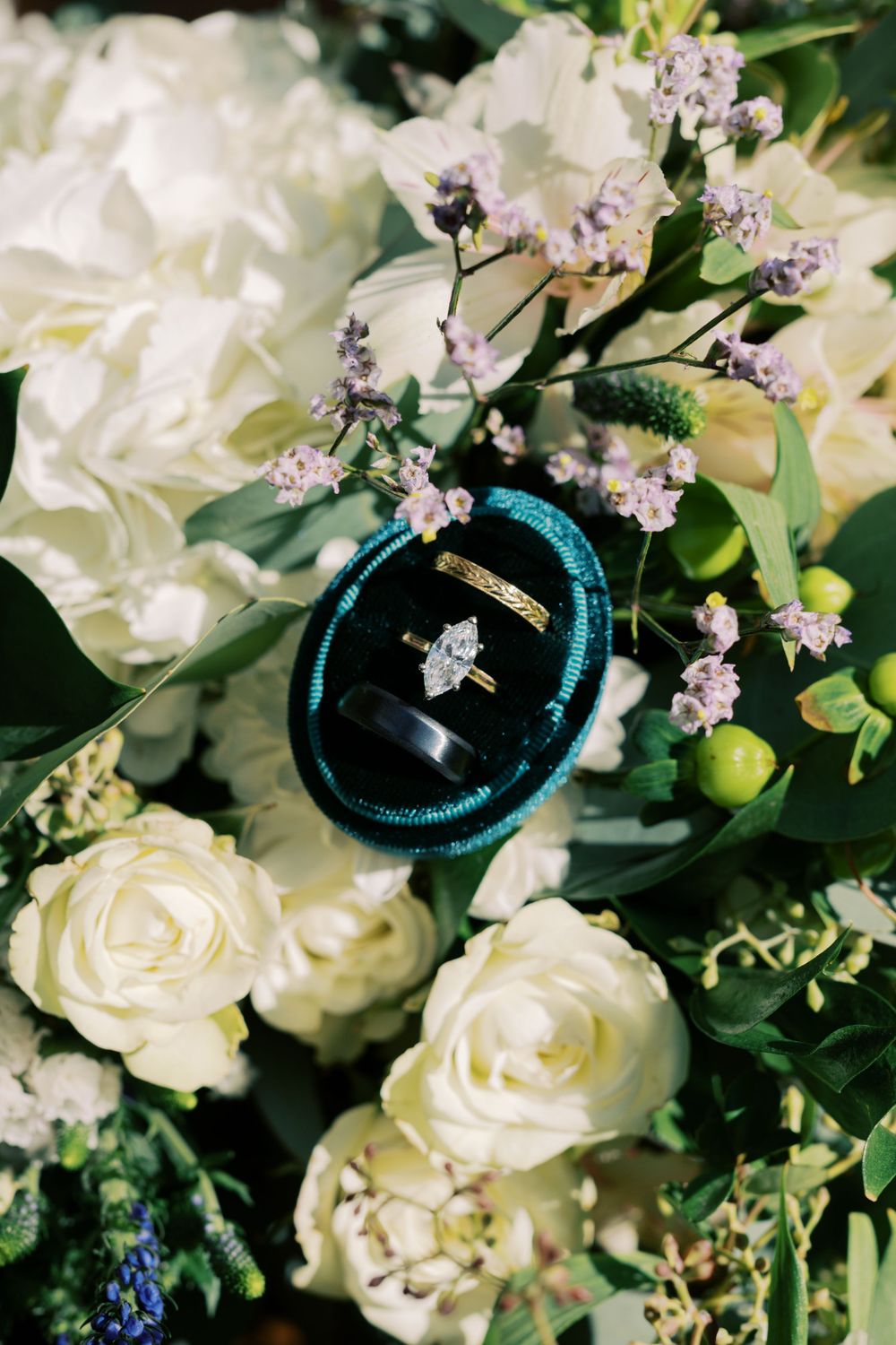 Close-up of wedding rings displayed on turquoise velvet cushion surrounded by white roses and delicate floral arrangements.