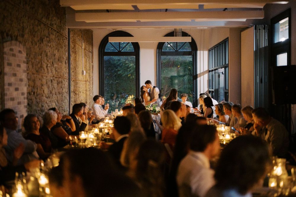 Candlelit dinner reception in stone-walled venue with guests seated at long tables under arched windows.