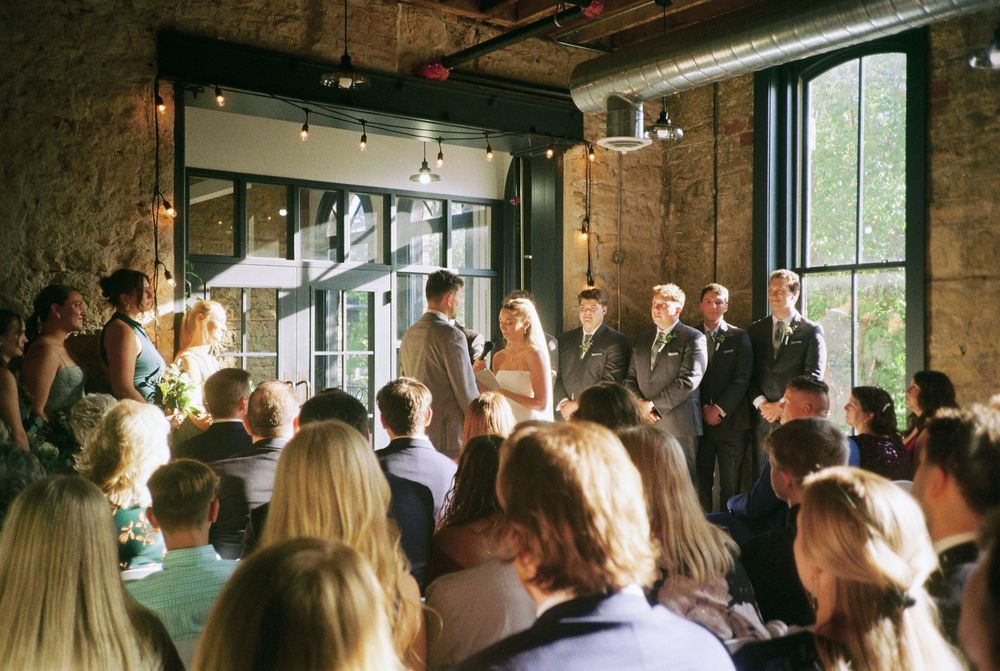 A crowd of wedding guests gathered in a rustic brick venue with large windows and natural lighting.