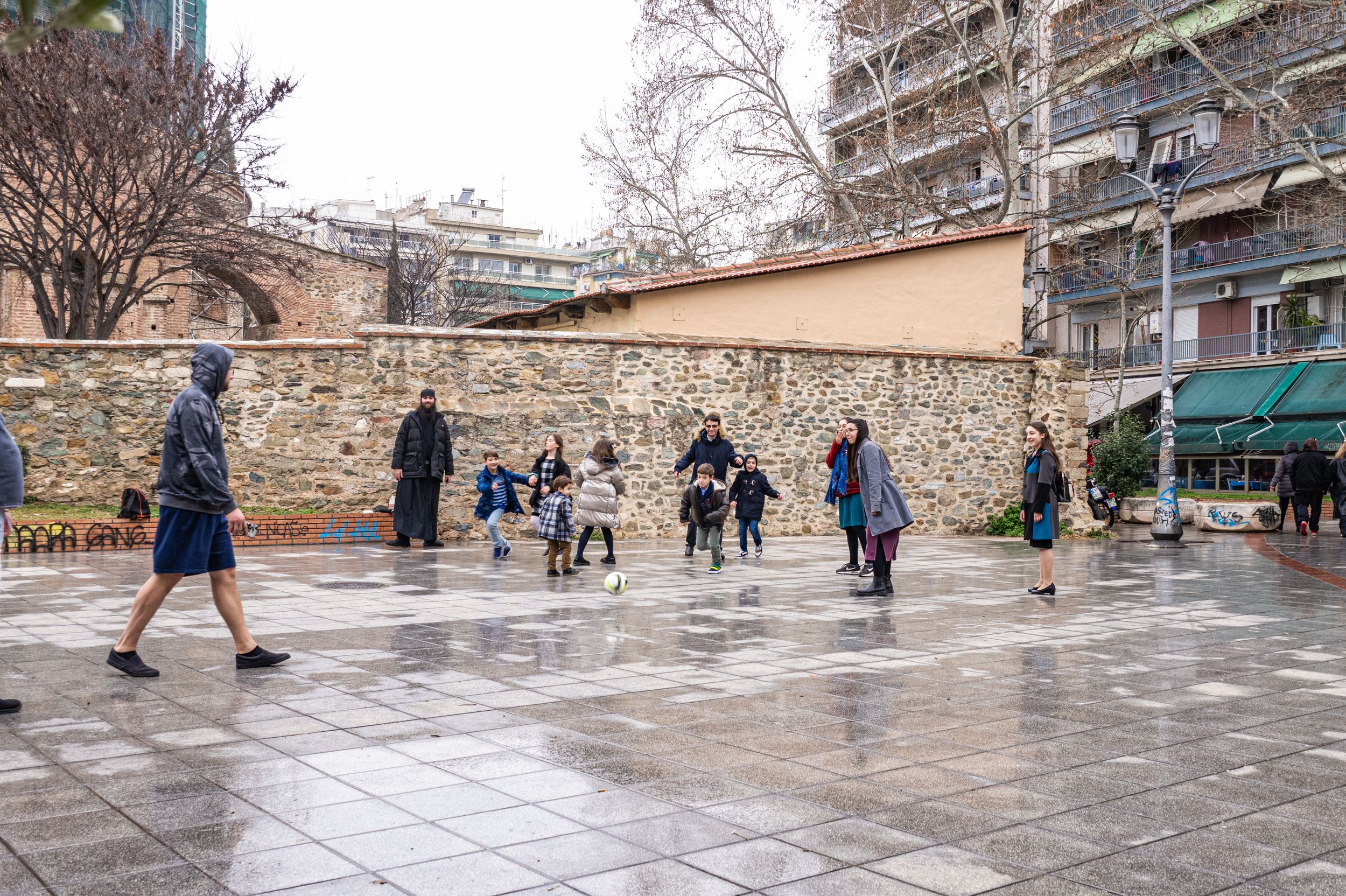 A bustling city plaza with people walking and standing on a wet, paved surface surrounded by buildings.