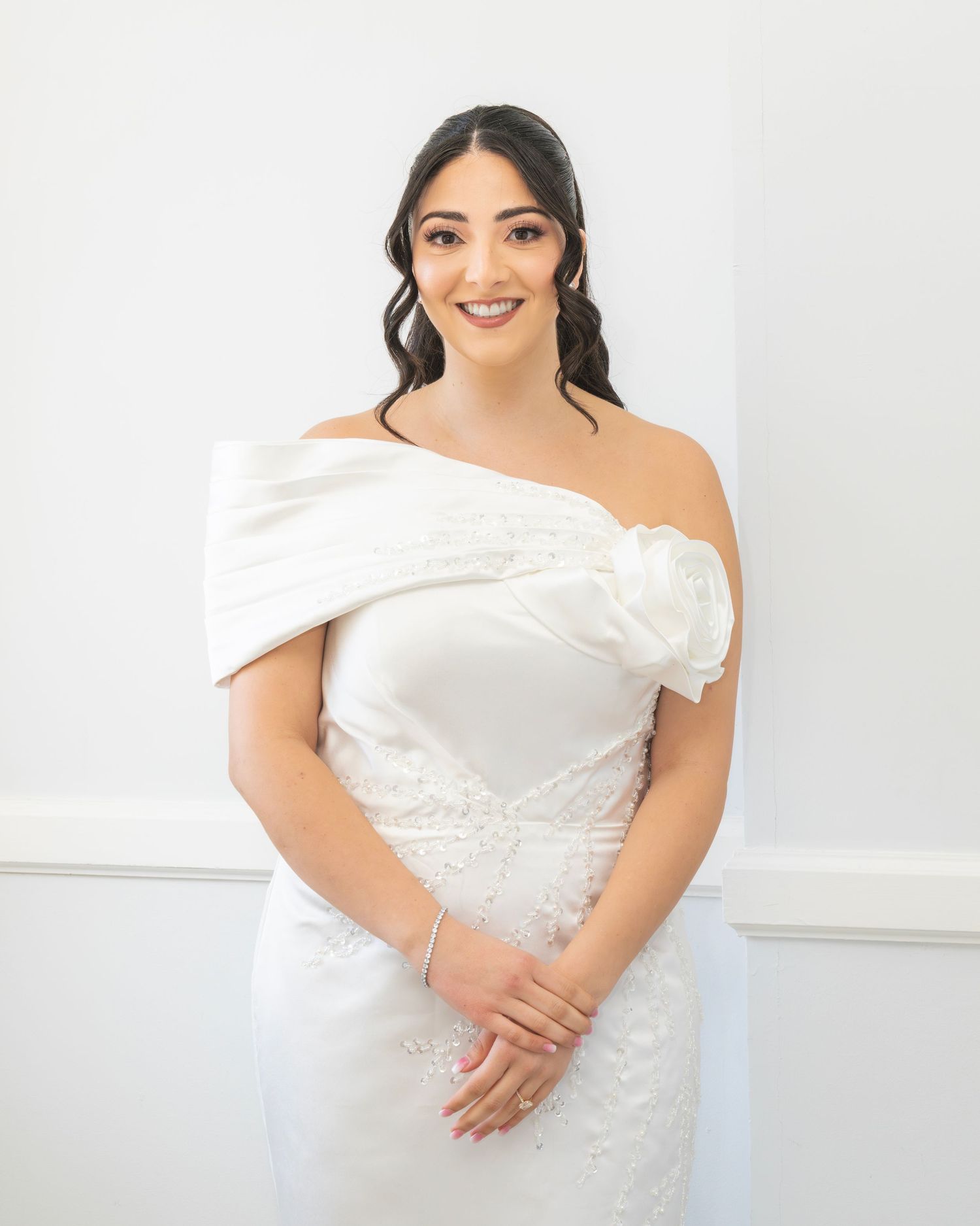 Smiling bride in an elegant off-shoulder white wedding gown with floral detail, posing indoors with hands clasped.