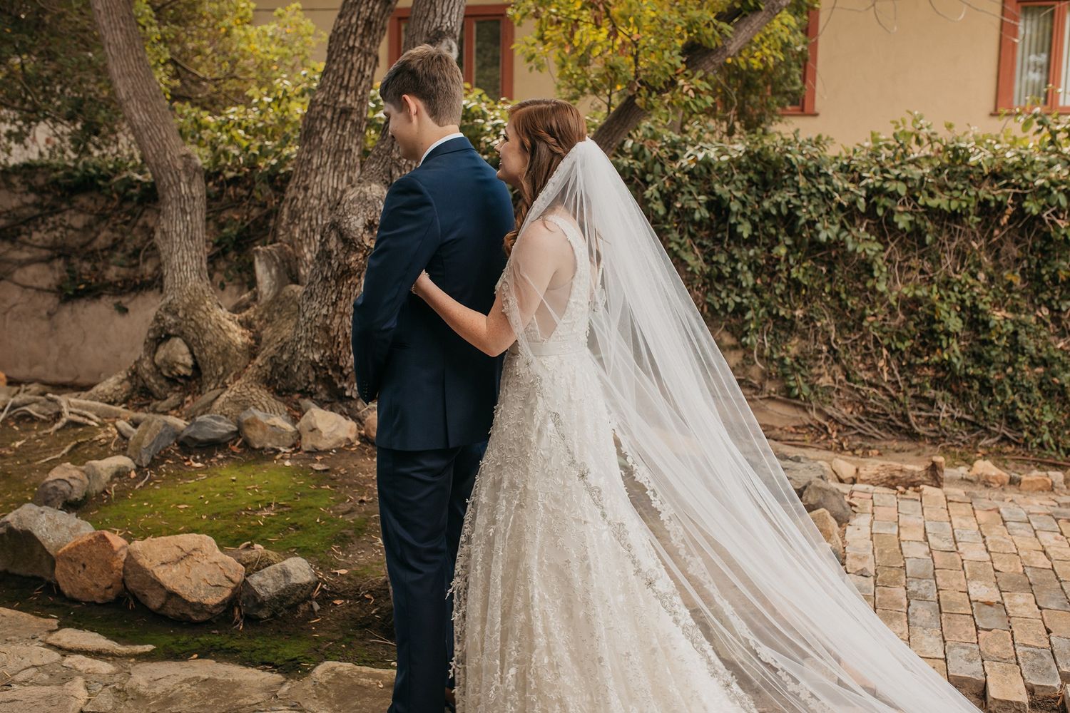 Bride and groom share an intimate moment outdoors, bride wearing a lace gown with long veil, groom in navy suit at Rancho Buena Vista Adobe.