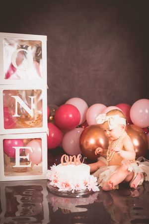 Baby girl celebrates first birthday with white cake, ONE letter boxes, and pink and gold balloons in a studio photo.