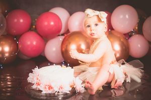 Baby girl in white tutu sits beside first birthday cake surrounded by pink and gold balloons.