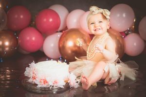 Smiling baby girl in tutu and pearl necklace celebrates first birthday with white cake and pink balloons backdrop.