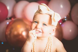 Smiling baby girl with a bow headband eating cake at a first birthday party with pink and gold balloons.