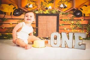 Baby's first birthday cake smash photo with bee theme, ONE sign, honeycomb backdrop, and fairy lights.