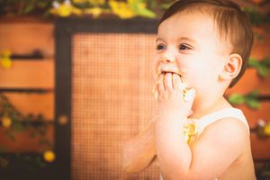 A happy baby in a white outfit joyfully eating cake with both hands outdoors in a warm, sunlit setting.