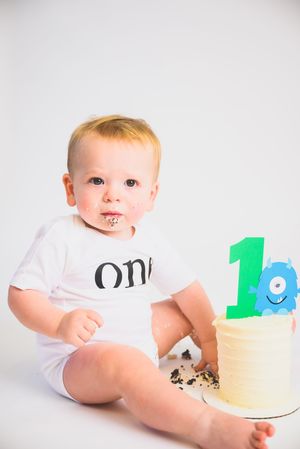 Baby boy in white onesie celebrating first birthday with smash cake topped with number one monster topper.