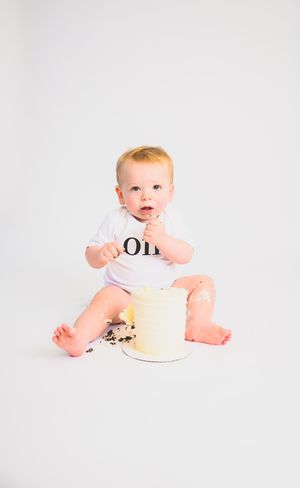 Baby in ONE onesie smashing first birthday cake on white background.
