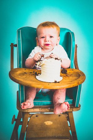 Messy baby in vintage high chair smashing first birthday cake with chocolate frosting on teal background.