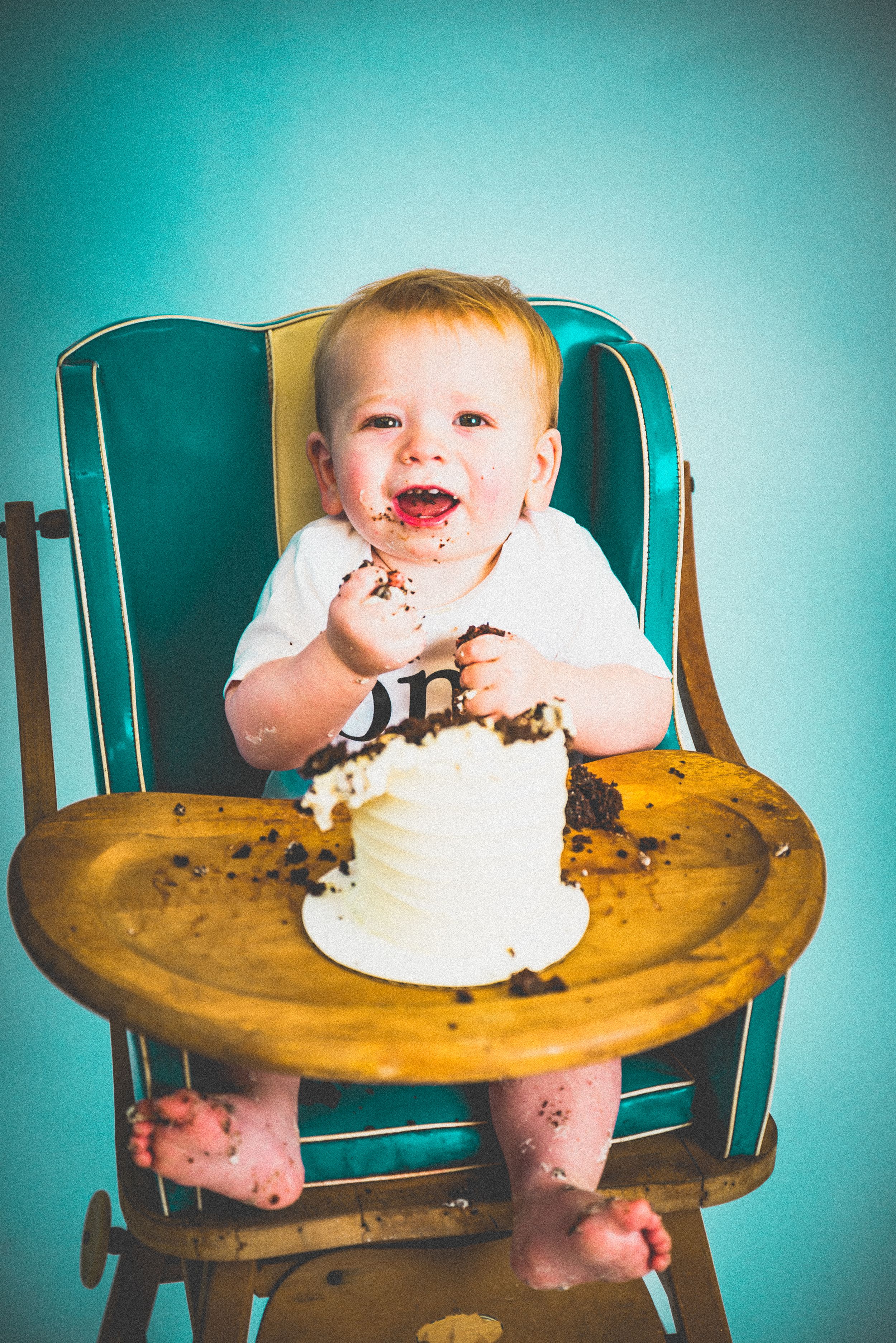 Happy baby in vintage high chair smashing first birthday cake, covered in frosting and crumbs.