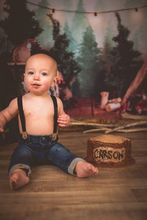 Baby boy named Carson sits on floor in jeans and suspenders beside a rustic cake for his first birthday.