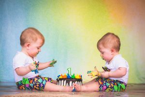 Twin babies in colorful pants smash a first birthday cake together, hands covered in paint.