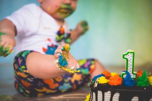 Baby with colorful painted hands smashes first birthday cake with number one candle on floor.
