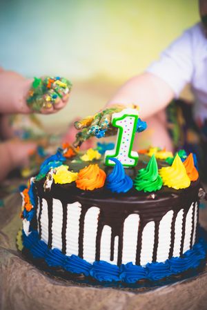 Colorful first birthday cake with chocolate drip, blue frosting, and a number 1 candle being smashed by a baby.