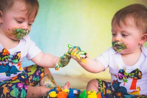Twin babies in dinosaur outfits share colorful cake smash with painted faces and hands touching.