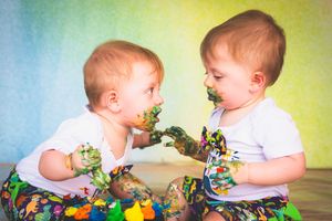 Twin babies with paint-covered hands and faces laugh together during a first birthday smash cake.