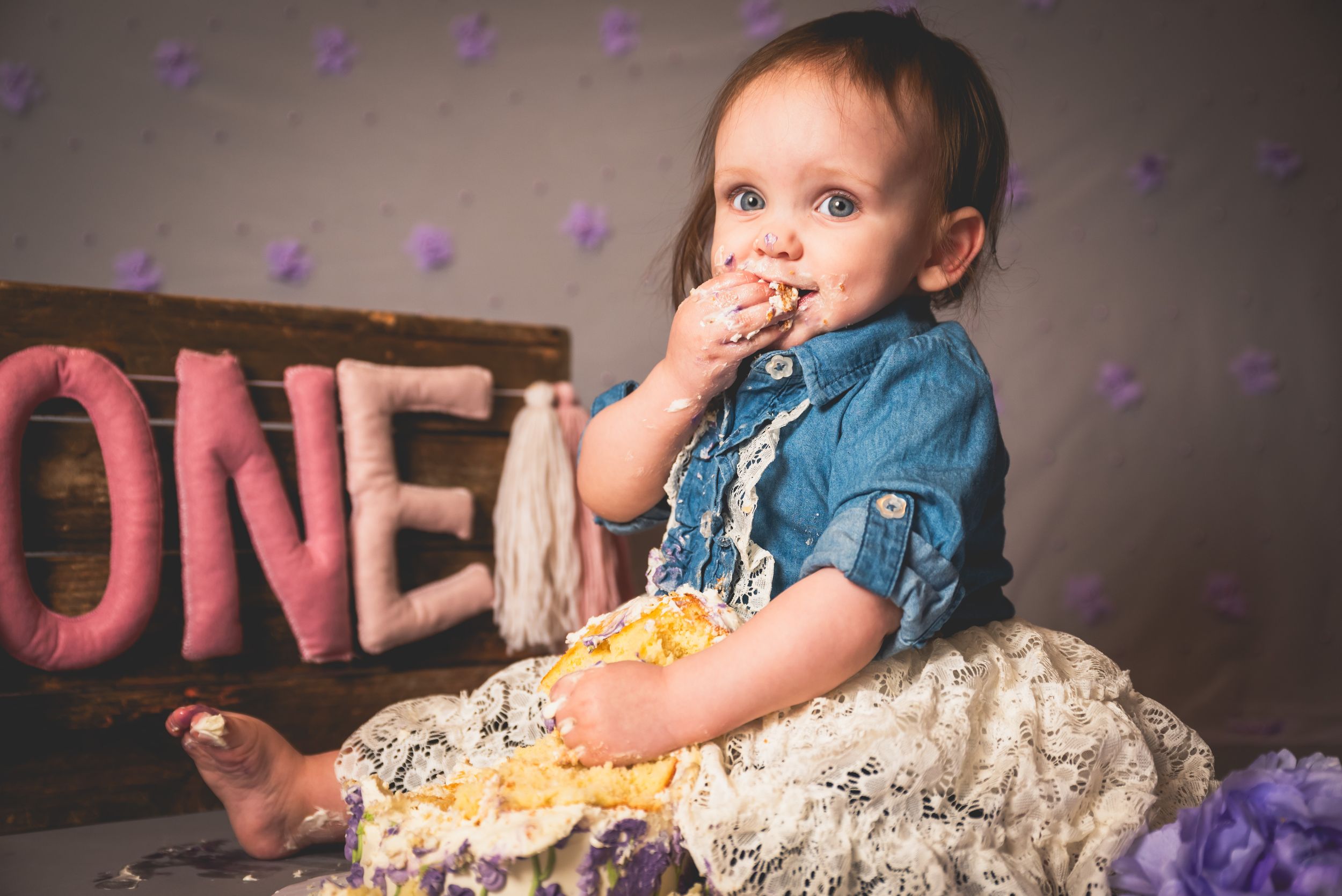 Baby girl in denim shirt eating smash cake during first birthday photo session with ONE sign.