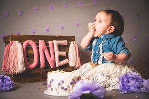 Baby enjoying first birthday cake smash with ONE sign, purple flowers, and white cake on gray background.