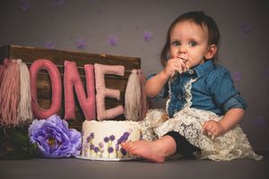 Baby girl celebrating first birthday smashing a floral cake beside a wooden ONE sign in a studio photo.