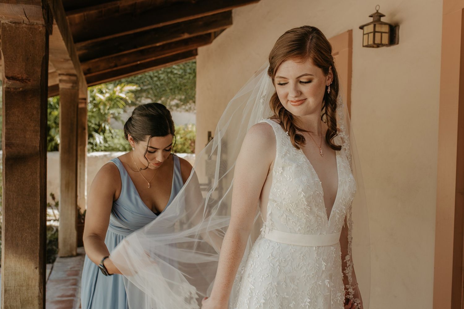 Bridesmaid in blue dress adjusting bride's white lace wedding veil outdoors at Rancho Buena Vista Adobe.