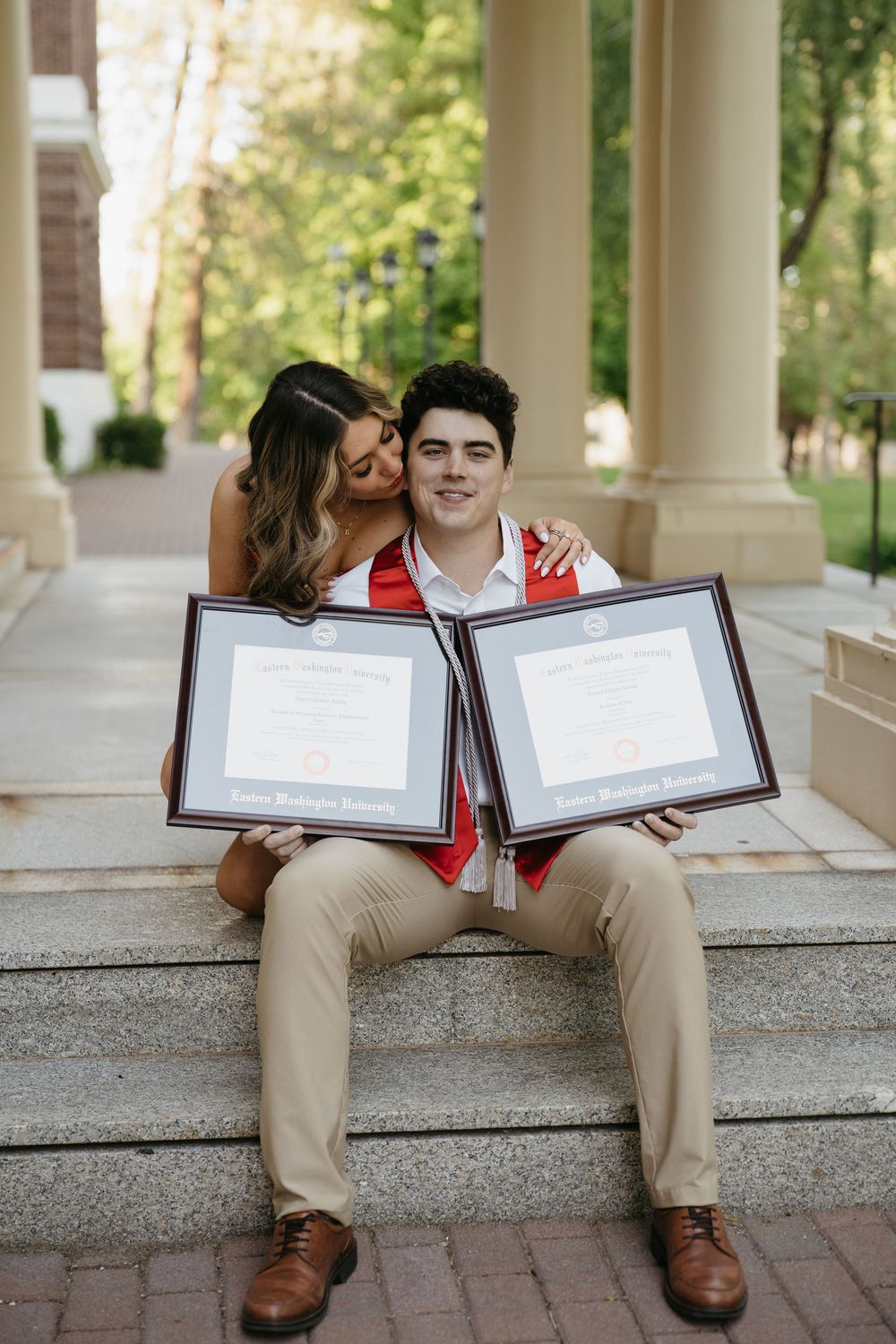 Two graduates holding diplomas on stone steps, smiling and celebrating their academic achievement outdoors.
