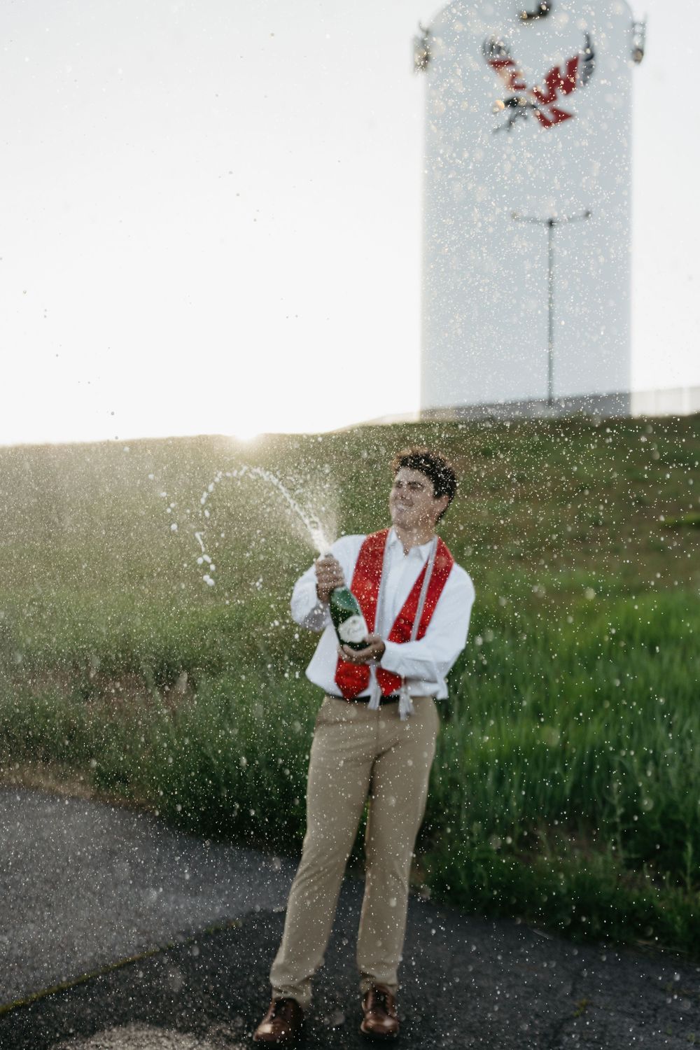 Young man in traditional attire joyfully spraying champagne outdoors at sunset near a decorated monument.