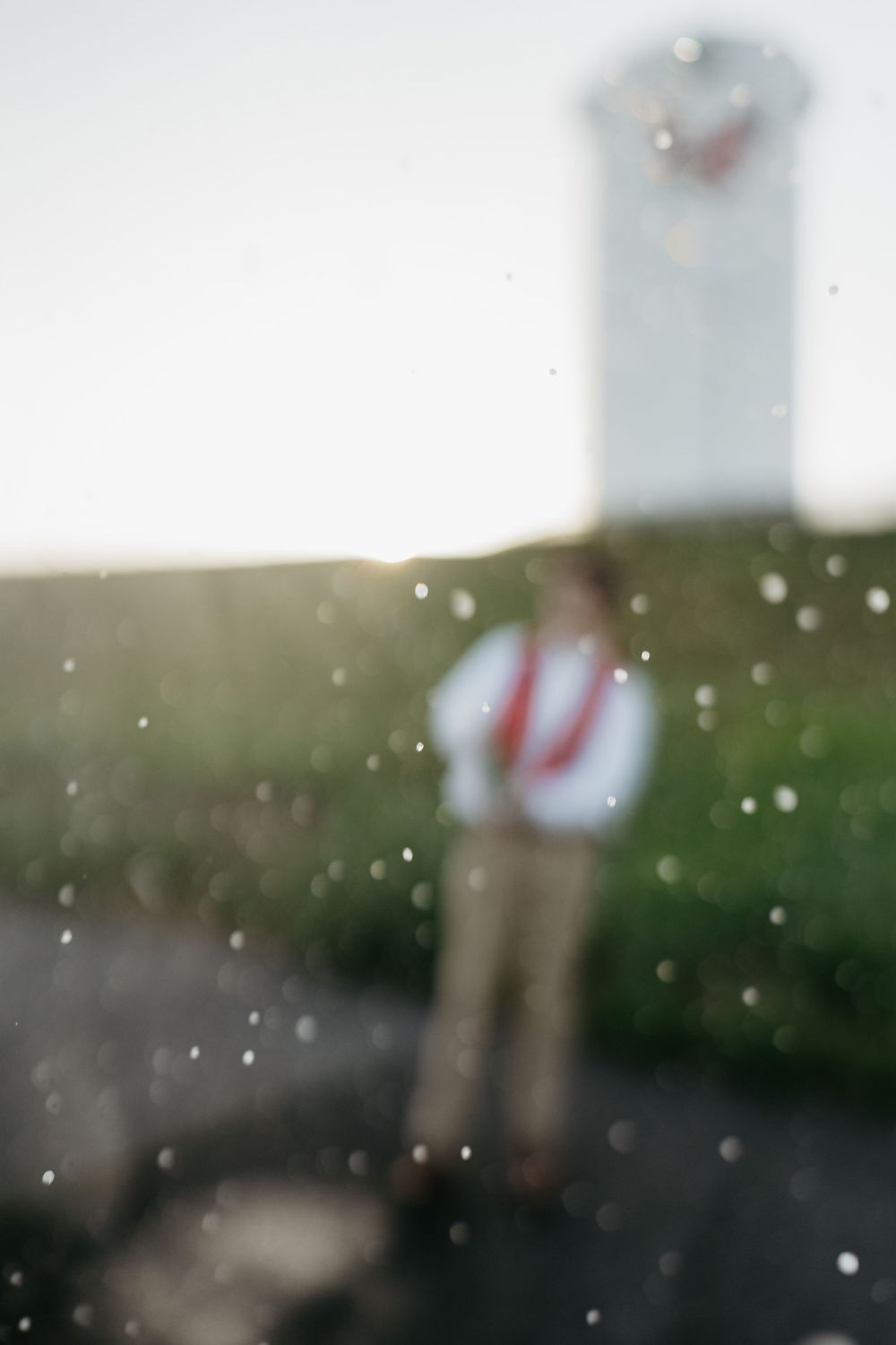 Blurred figure in white shirt and red tie stands outdoors near a tower, viewed through rain-spotted glass.
