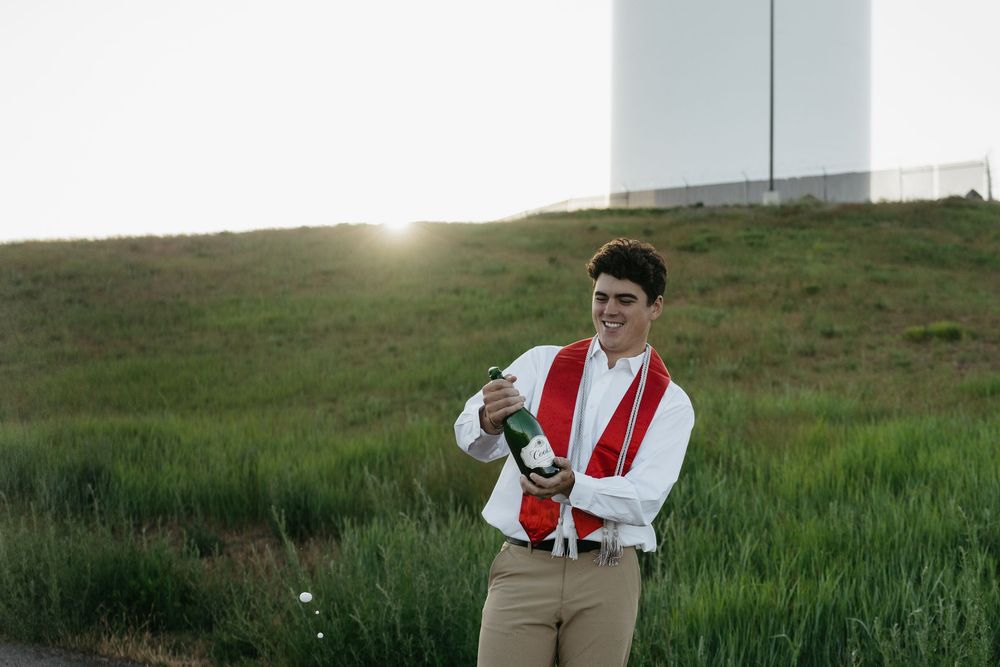 Young man in white shirt and red sash opening champagne bottle outdoors at sunset near green hillside.