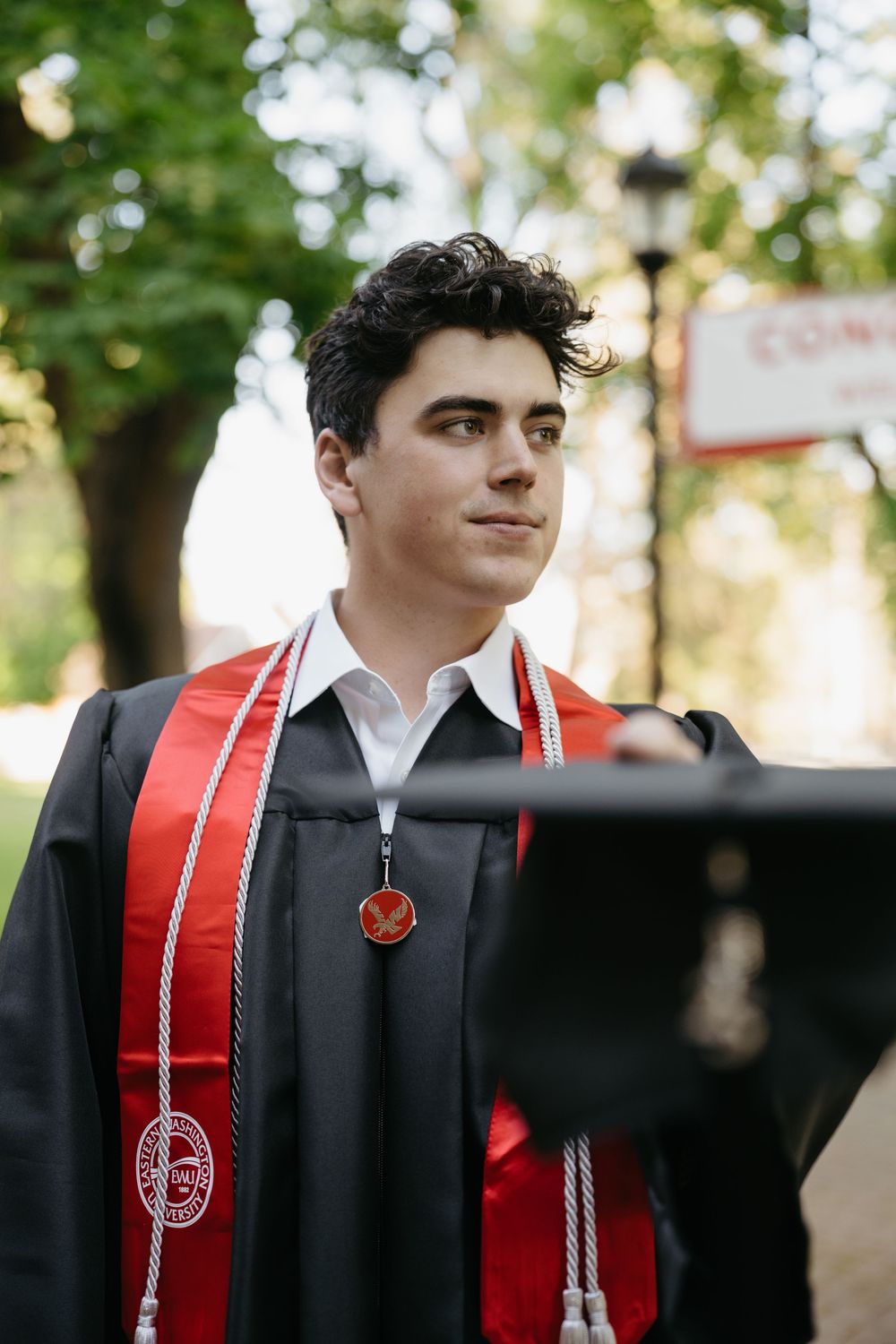 Young man in black graduation gown with red stole and honor cords poses outdoors on campus.