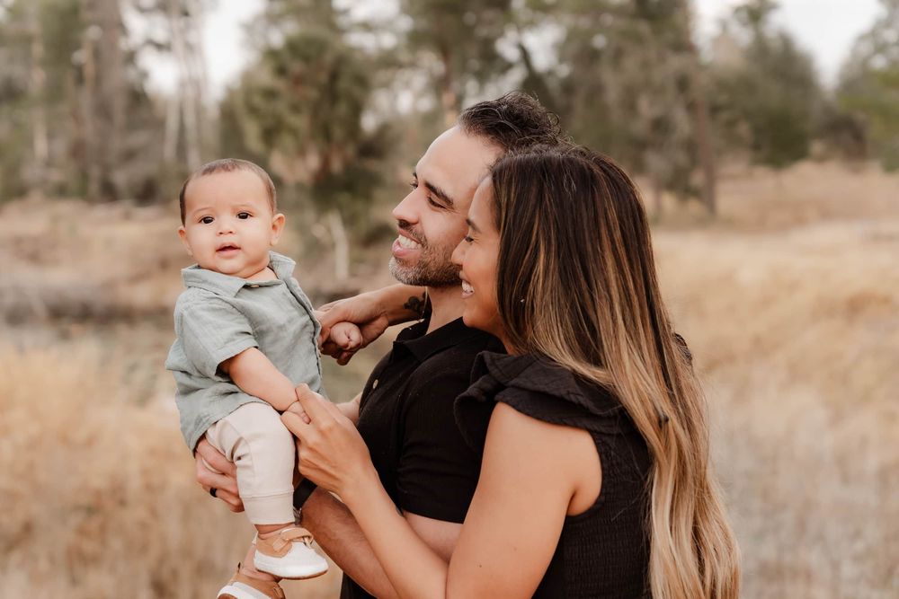 A parent holds a baby in a gray outfit while standing in a golden field during sunset.