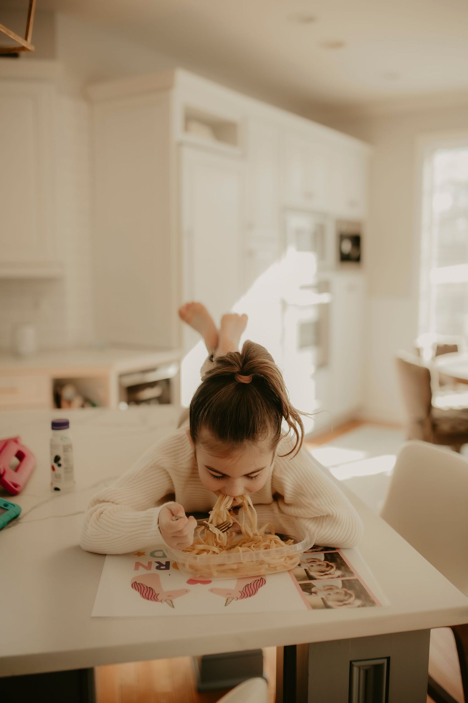 A child leans over a white table while drawing or coloring in a bright, sunlit room.