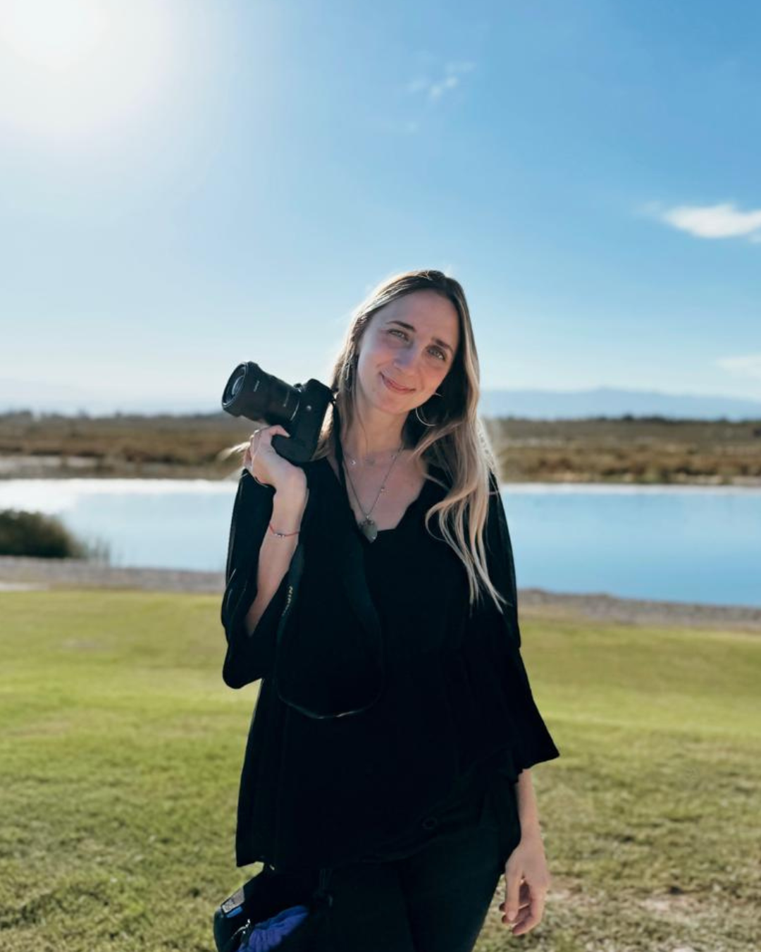 Blonde female photographer holding a camera outdoors near a lake on a sunny day.