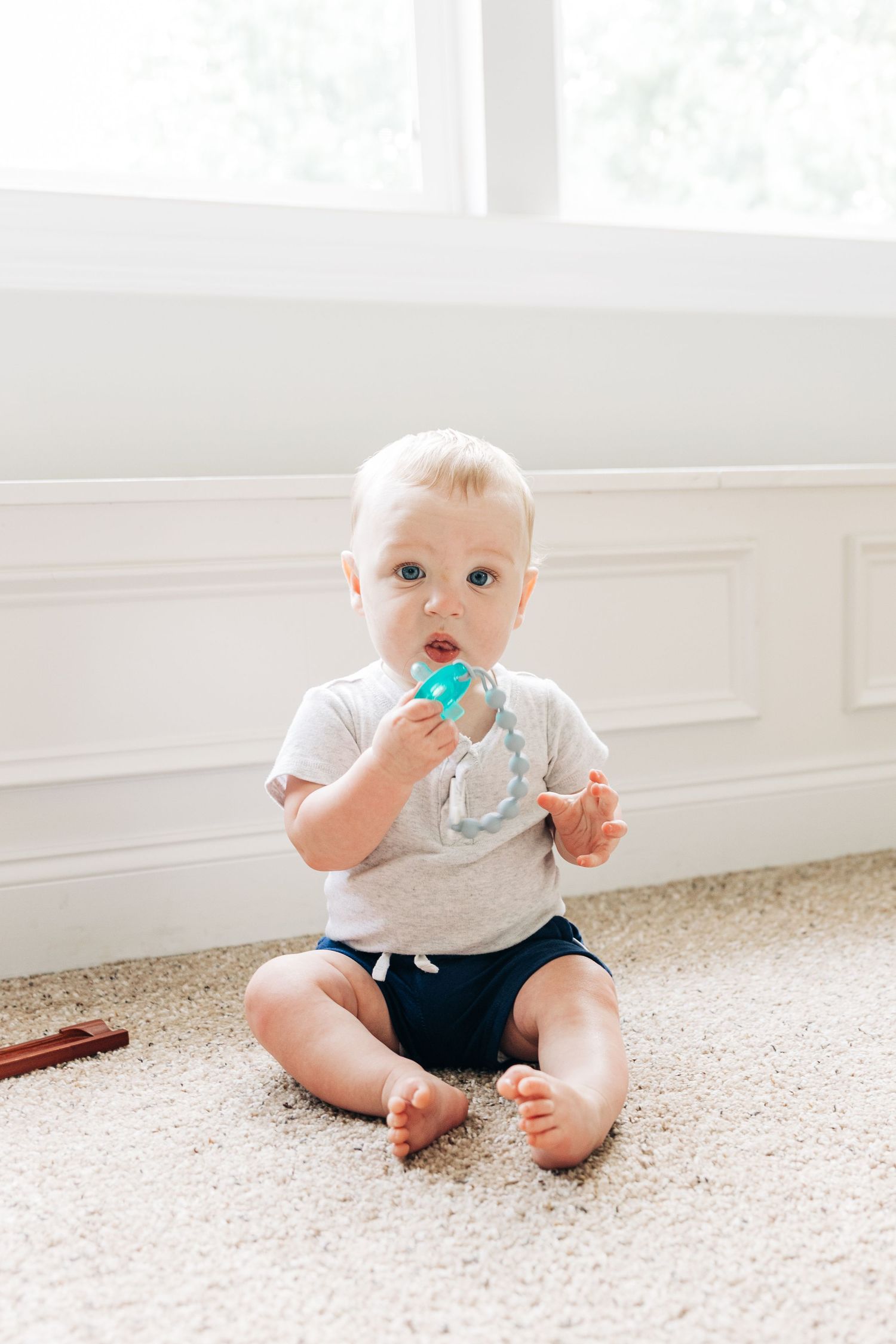 Baby in white shirt and navy shorts sits on beige carpet with pacifier while crawling.