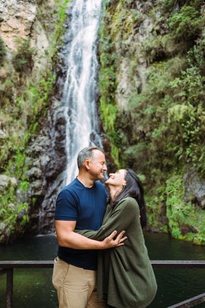 A couple shares a romantic moment in front of a scenic waterfall surrounded by lush greenery.