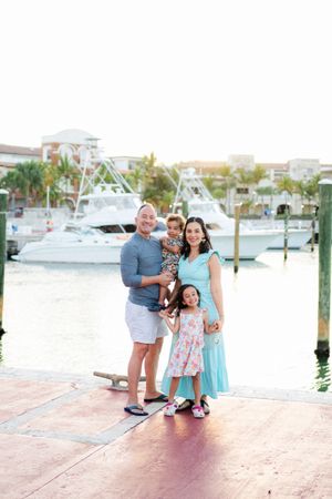 A family enjoys a sunset moment together on a marina dock with boats and palm trees in the background.