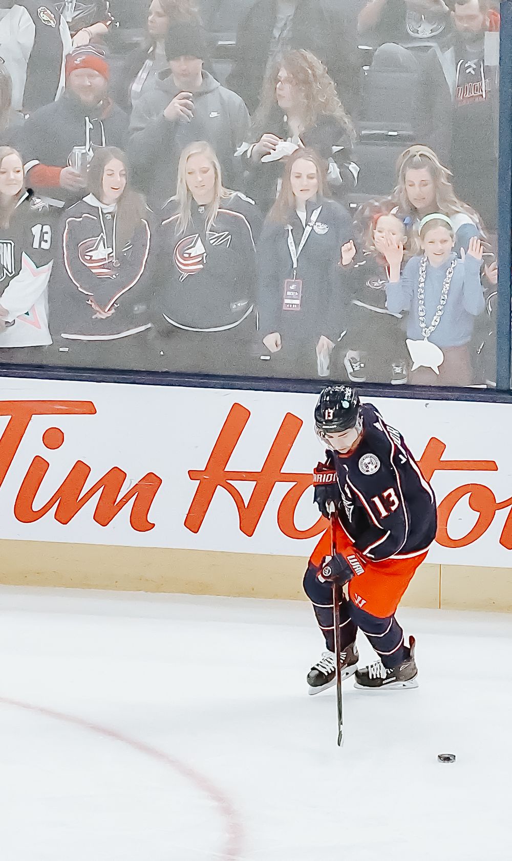 Hockey player in dark jersey skates on ice rink while spectators watch through glass barrier during game.