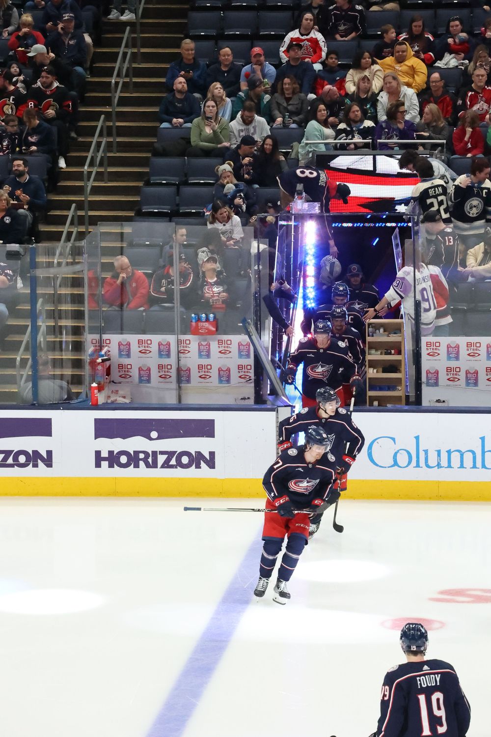 Hockey players skate on ice during a professional game with advertisements and crowd visible in the background.