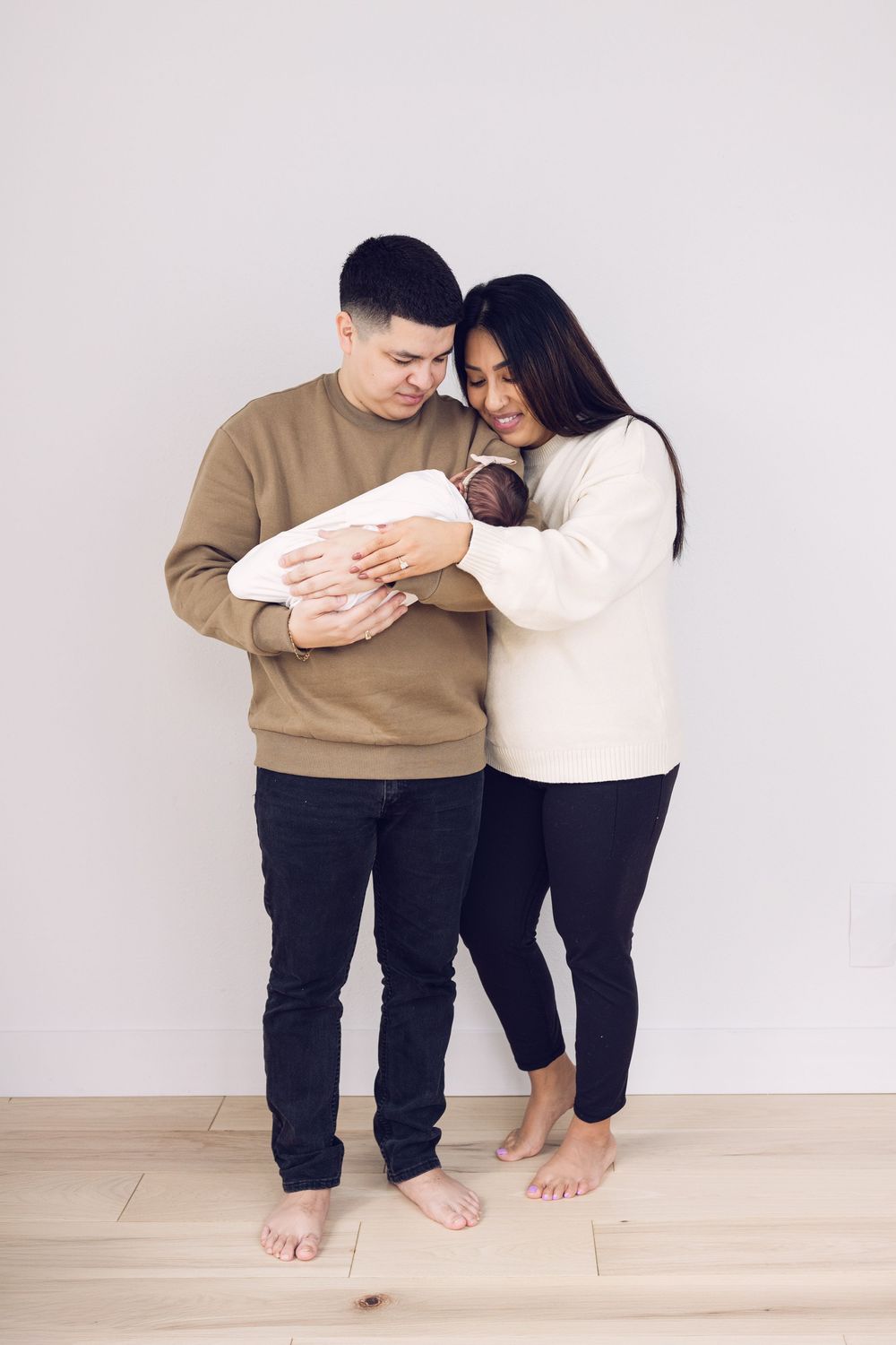 Young couple standing against white wall smiling while holding their swaddled newborn baby.