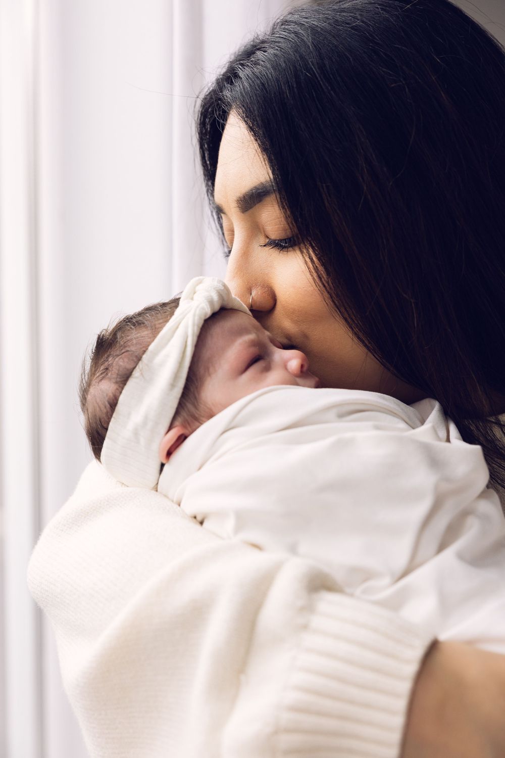 Dark-haired mother gently kissing her swaddled newborn baby near a bright window.