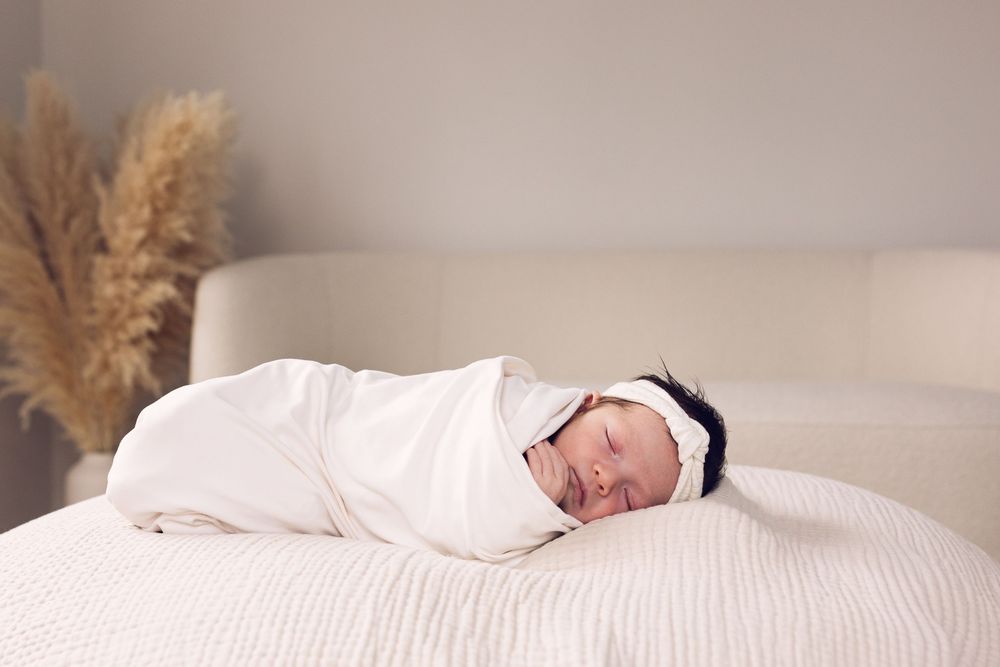 Swaddled newborn baby sleeping peacefully on a white bed with a dark headband.