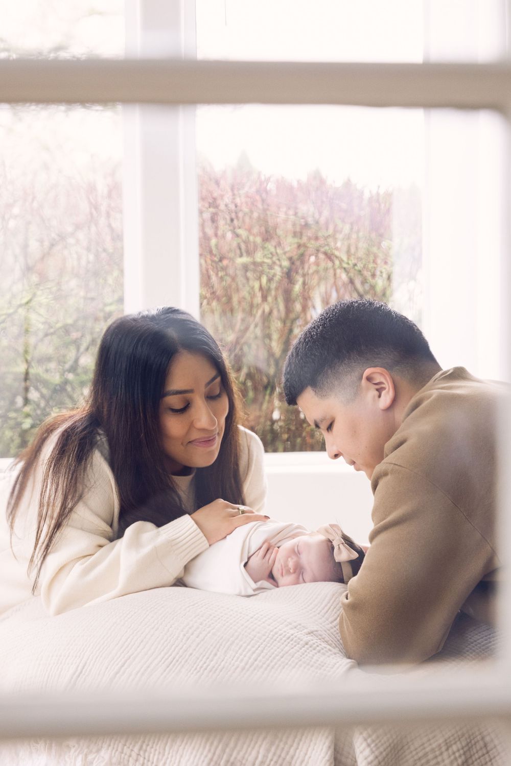 New parents tenderly gazing at their newborn baby by a sunlit window.