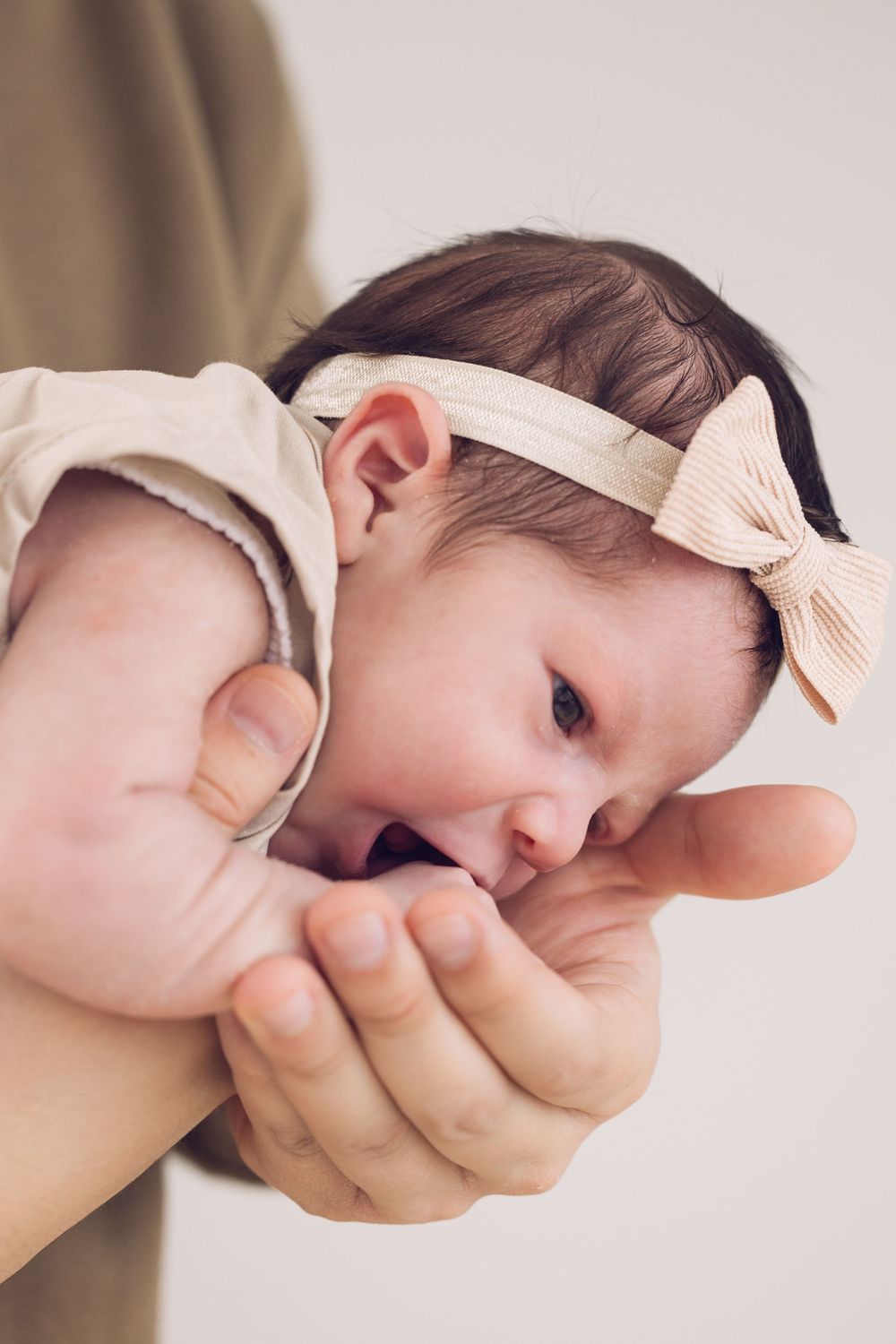 Newborn baby girl with bow headband being held in father's hands, sucking thumb.