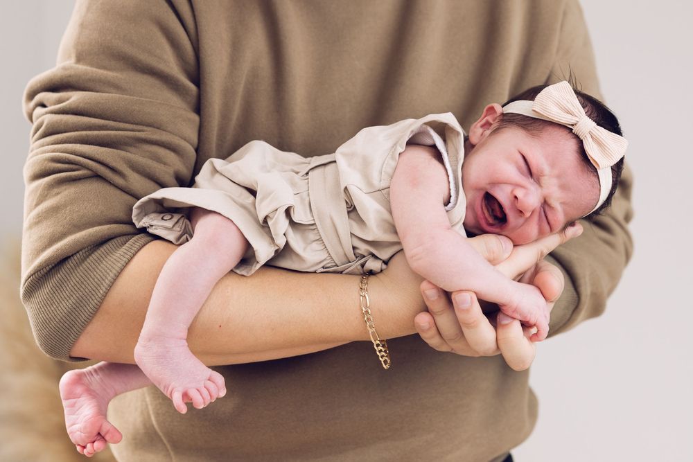 Crying newborn baby in beige outfit and bow headband being held in adult hands.