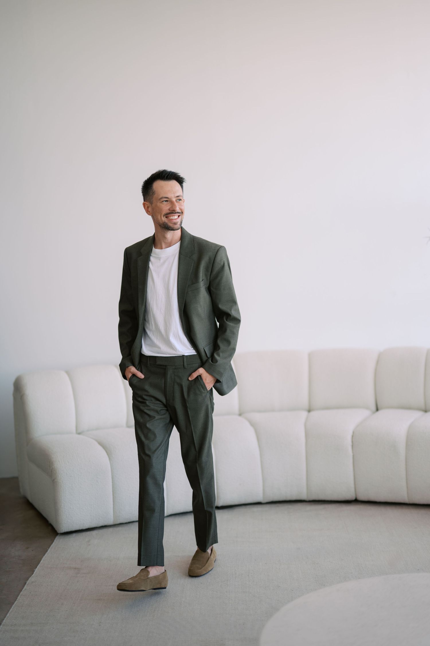 A person wearing an olive green suit and white t-shirt stands casually against a white wall near a curved sofa.